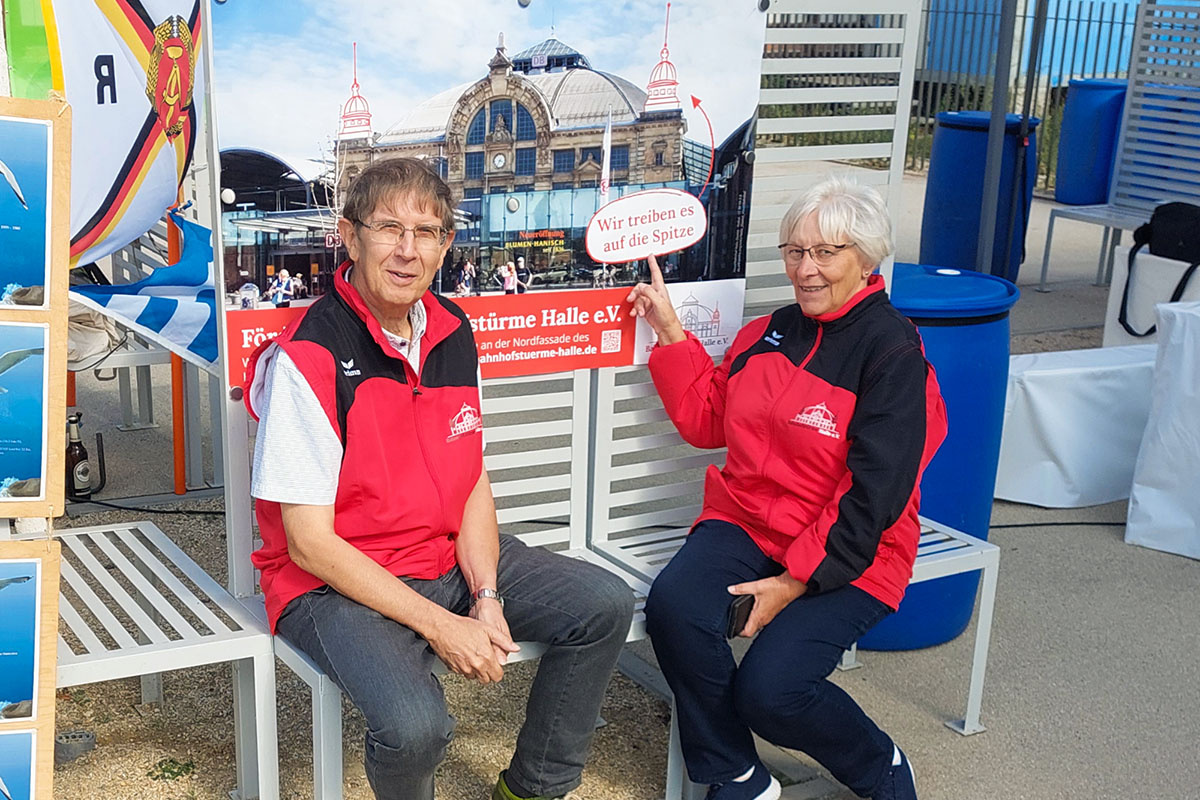 Zwei Vereinsmitglieder sitzen beim Salzfest and der Saline auf einer Bank. Ein Vereinsmitglied zeigt auf ein Plakat im Hintergrund auf dem "Wir treiben es auf die Spitze" steht.